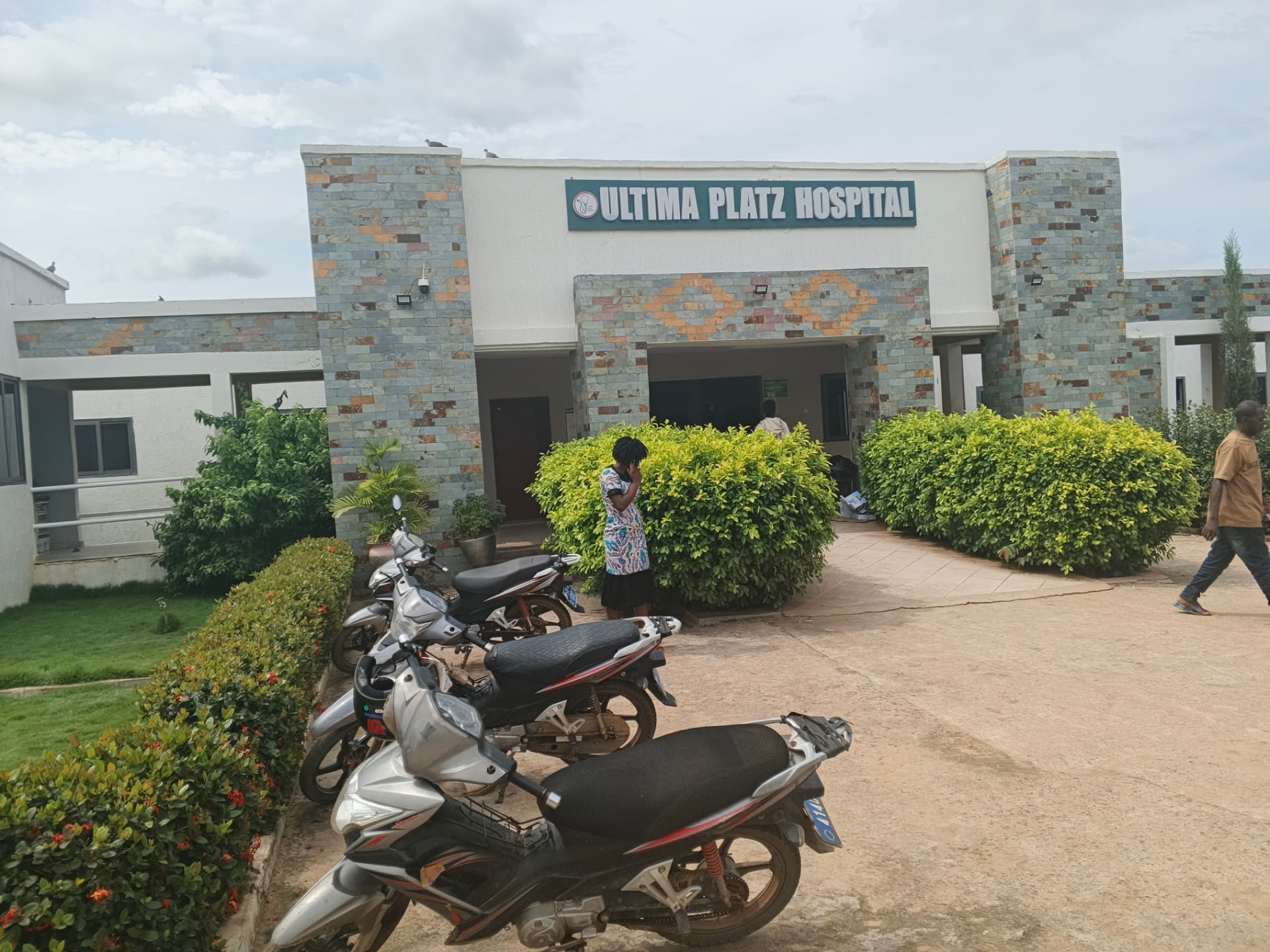 The main entrance of Ultima Platz Hospital, a single-storey building clad in multicoloured stone with the hospital name on a teal sign above the doorway.
